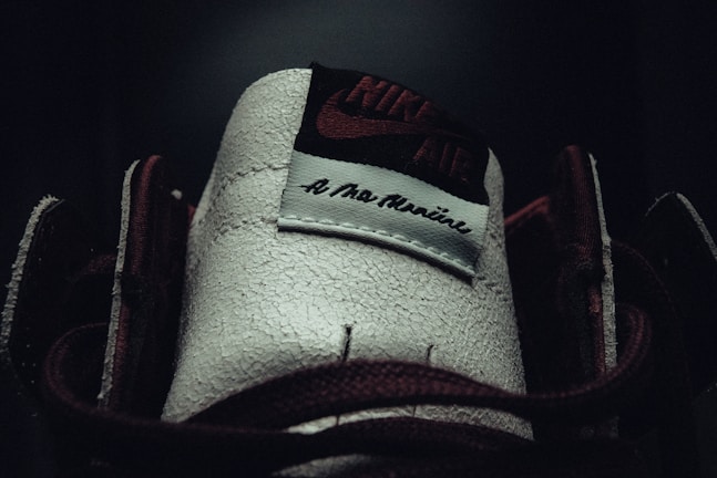 A close-up of a shoe tongue featuring the Nike Air logo and the phrase 'A Ma Maniére' on a textured surface. The lighting is dim, highlighting the contrast between the dark and light areas.