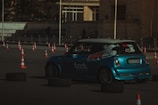A blue racing car navigates a course marked by orange traffic cones and black tires. The car has visible branding and decals on the side and back. The setting appears to be an outdoor paved area with barriers in the background.