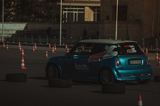 A blue racing car navigates a course marked by orange traffic cones and black tires. The car has visible branding and decals on the side and back. The setting appears to be an outdoor paved area with barriers in the background.