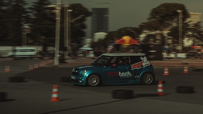 A blue racing car navigates a course marked by orange traffic cones. The scene depicts motion with the car slightly blurred, emphasizing speed. The background shows a few trees and a tall building under a cloudy sky.