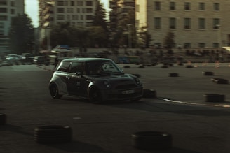 A compact car with a sleek, dark exterior is navigating a course marked with tires and cones during what appears to be an auto race or drift event. The background includes a few buildings and a crowd of spectators.