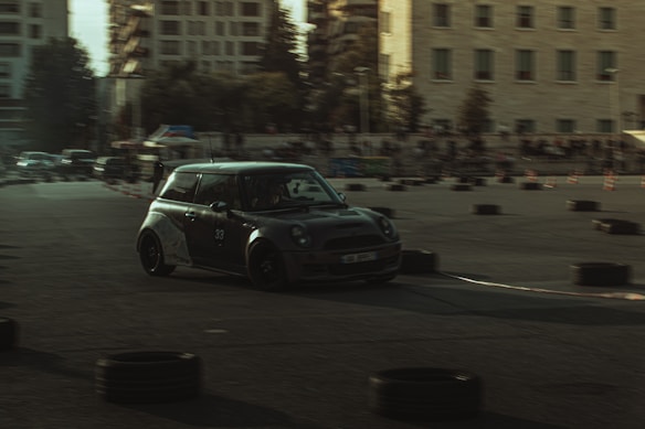A compact car with a sleek, dark exterior is navigating a course marked with tires and cones during what appears to be an auto race or drift event. The background includes a few buildings and a crowd of spectators.