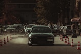 A car performing a drift maneuver on a road lined with traffic cones, surrounded by spectators and other parked vehicles. Dust kicks up behind the car, indicating speed and movement. The background shows a mix of urban elements, including trees and a building, with people casually observing the event.