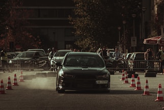 A car performing a drift maneuver on a road lined with traffic cones, surrounded by spectators and other parked vehicles. Dust kicks up behind the car, indicating speed and movement. The background shows a mix of urban elements, including trees and a building, with people casually observing the event.