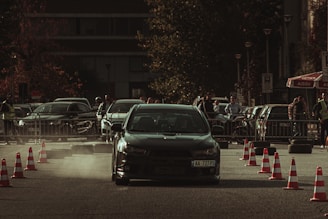 A car performing a drift maneuver on a road lined with traffic cones, surrounded by spectators and other parked vehicles. Dust kicks up behind the car, indicating speed and movement. The background shows a mix of urban elements, including trees and a building, with people casually observing the event.