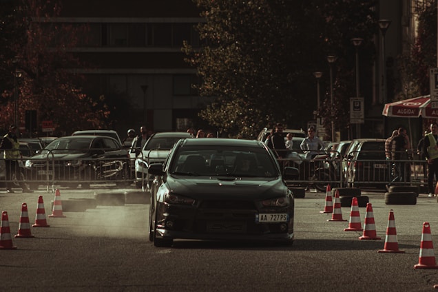 A car performing a drift maneuver on a road lined with traffic cones, surrounded by spectators and other parked vehicles. Dust kicks up behind the car, indicating speed and movement. The background shows a mix of urban elements, including trees and a building, with people casually observing the event.