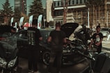A gathering of people around several cars and motorcycles in an outdoor setting with a modern building in the background. Some of the cars have their hoods open, indicating a possible car show or exhibition. Various flags are displayed along with tents, suggesting an organized event. People are interacting with the vehicles or each other, and the scene takes place on a sunny day.