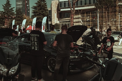 A gathering of people around several cars and motorcycles in an outdoor setting with a modern building in the background. Some of the cars have their hoods open, indicating a possible car show or exhibition. Various flags are displayed along with tents, suggesting an organized event. People are interacting with the vehicles or each other, and the scene takes place on a sunny day.