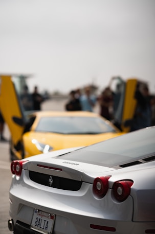 A close-up view of a silver Ferrari with its back facing the viewer, featuring distinctive tail lights and a California license plate. In the background, a blurred yellow sports car with upward-opening doors can be seen, along with several people standing around.