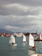 Several small sailboats with young sailors take part in a sailing activity on a body of water. The background features a cityscape with multiple large ships docked at a harbor. The sky is overcast, indicating cloudy weather.
