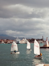 Several small sailboats with young sailors take part in a sailing activity on a body of water. The background features a cityscape with multiple large ships docked at a harbor. The sky is overcast, indicating cloudy weather.