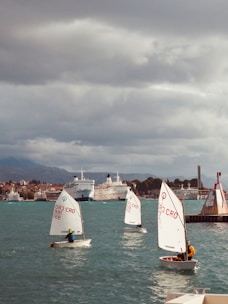 Several small sailboats with young sailors take part in a sailing activity on a body of water. The background features a cityscape with multiple large ships docked at a harbor. The sky is overcast, indicating cloudy weather.