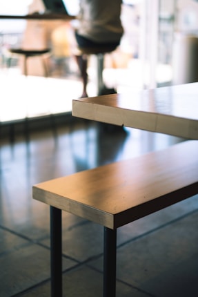 A wooden table with metal legs is prominently featured in the foreground, set in a cafe environment. In the background, there are blurred figures, possibly a person sitting by the window with a laptop, underlining a casual and relaxed atmosphere.
