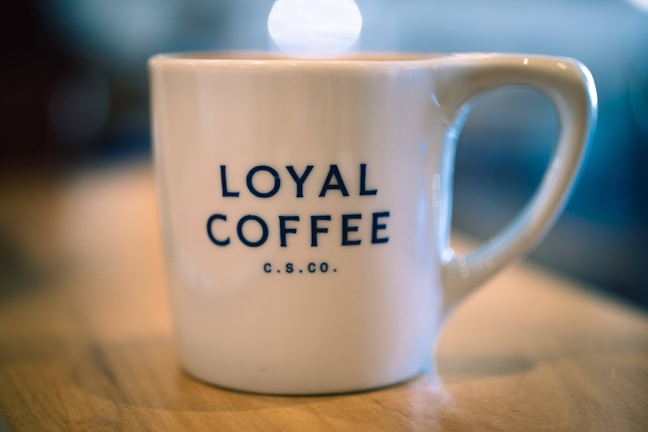 A white ceramic coffee mug with the words 'Loyal Coffee C.S.CO.' printed in bold, dark blue letters sits on a wooden surface. The background is softly blurred, highlighting the mug as the focal point.