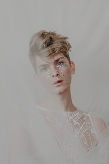 A person with short, styled hair and rhinestones adorning their face wears an intricately patterned and sheer fabric garment. The background is soft and blurred, adding a dreamy effect to the image.