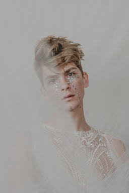 A person with short, styled hair and rhinestones adorning their face wears an intricately patterned and sheer fabric garment. The background is soft and blurred, adding a dreamy effect to the image.