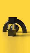 A minimalist composition featuring geometric shapes arranged against a bright yellow background. Black cubes, a transparent block, and a spherical shape are arranged in front of two large curved black cylinders. The scene is clean and smooth with stark contrasts between the black objects and the vibrant yellow surface.