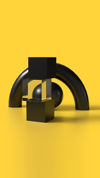 A minimalist composition featuring geometric shapes arranged against a bright yellow background. Black cubes, a transparent block, and a spherical shape are arranged in front of two large curved black cylinders. The scene is clean and smooth with stark contrasts between the black objects and the vibrant yellow surface.