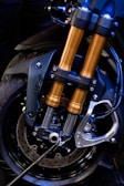 A close-up view of a motorcycle front suspension fork and brake system, highlighting the metallic structure with black and gold components. The intricate design showcases the engineering and craftsmanship involved, with visible screws, bolts, and brake discs.