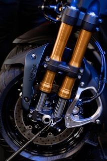 A close-up view of a motorcycle front suspension fork and brake system, highlighting the metallic structure with black and gold components. The intricate design showcases the engineering and craftsmanship involved, with visible screws, bolts, and brake discs.