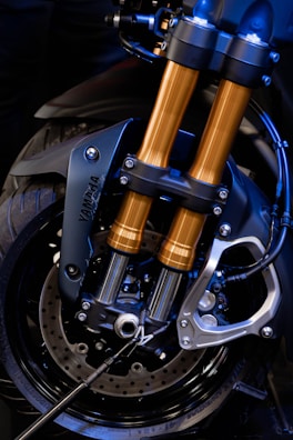 A close-up view of a motorcycle front suspension fork and brake system, highlighting the metallic structure with black and gold components. The intricate design showcases the engineering and craftsmanship involved, with visible screws, bolts, and brake discs.