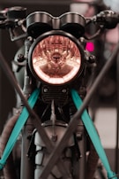 A close-up of a motorcycle front with a prominent headlight encased in a circular design. The motorcycle has distinctive handlebars and features teal straps securing it. It appears to be displayed or parked, with modern and rugged elements visible.