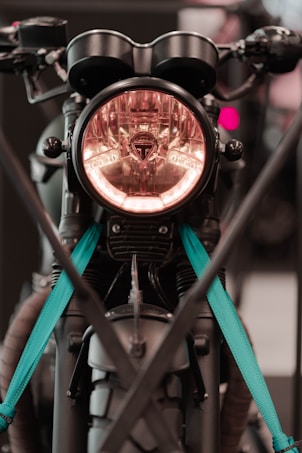 A close-up of a motorcycle front with a prominent headlight encased in a circular design. The motorcycle has distinctive handlebars and features teal straps securing it. It appears to be displayed or parked, with modern and rugged elements visible.