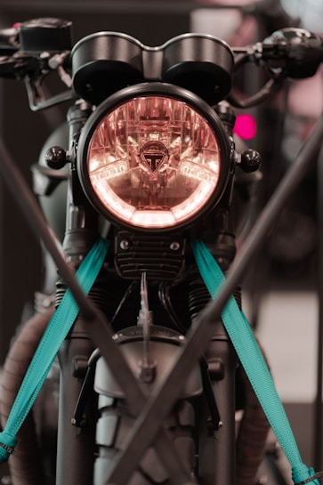 A close-up of a motorcycle front with a prominent headlight encased in a circular design. The motorcycle has distinctive handlebars and features teal straps securing it. It appears to be displayed or parked, with modern and rugged elements visible.