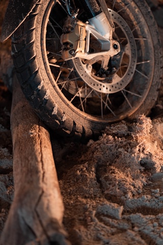 A motorcycle tire rests on a rough terrain composed of a mixture of sand and a wooden log. The tire is covered with dirt and the scene is illuminated with warm lighting, highlighting the texture of the sand and the ruggedness of the tire tread.