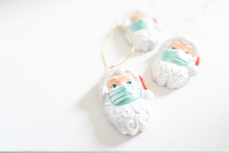 Three Santa Claus ornaments, each wearing a green face mask, are laid on a white surface. The ornaments are white with intricate beard details and have blue eyes. A golden string is attached to each for hanging.