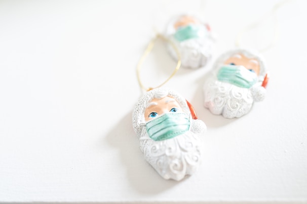 Three Santa Claus ornaments, each wearing a green face mask, are laid on a white surface. The ornaments are white with intricate beard details and have blue eyes. A golden string is attached to each for hanging.