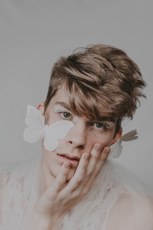 A person with soft features gently rests their face on their hand. Light-colored butterflies are delicately positioned on their face and ear, adding an ethereal touch. The overall appearance is dreamlike, with a soft focus and muted tones.