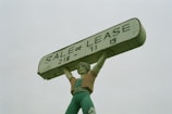 A large statue of a person holding up a rectangular sign above their head. The sign reads 'SALE OR LEASE' with a phone number. The statue is painted in shades of green, with a slightly weathered appearance.
