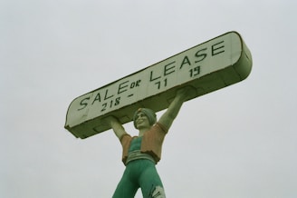 A large statue of a person holding up a rectangular sign above their head. The sign reads 'SALE OR LEASE' with a phone number. The statue is painted in shades of green, with a slightly weathered appearance.