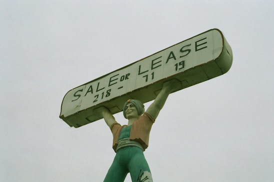 A large statue of a person holding up a rectangular sign above their head. The sign reads 'SALE OR LEASE' with a phone number. The statue is painted in shades of green, with a slightly weathered appearance.