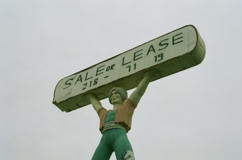 A large statue of a person holding up a rectangular sign above their head. The sign reads 'SALE OR LEASE' with a phone number. The statue is painted in shades of green, with a slightly weathered appearance.