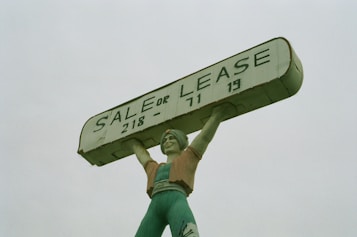 A large statue of a person holding up a rectangular sign above their head. The sign reads 'SALE OR LEASE' with a phone number. The statue is painted in shades of green, with a slightly weathered appearance.