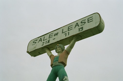 A large statue of a person holding up a rectangular sign above their head. The sign reads 'SALE OR LEASE' with a phone number. The statue is painted in shades of green, with a slightly weathered appearance.