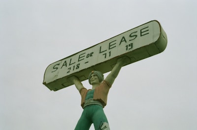 A large statue of a person holding up a rectangular sign above their head. The sign reads 'SALE OR LEASE' with a phone number. The statue is painted in shades of green, with a slightly weathered appearance.