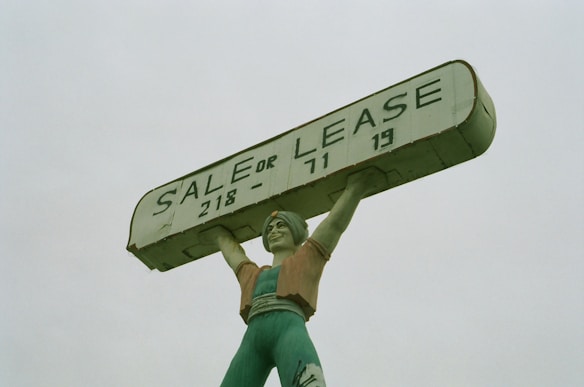 A large statue of a person holding up a rectangular sign above their head. The sign reads 'SALE OR LEASE' with a phone number. The statue is painted in shades of green, with a slightly weathered appearance.