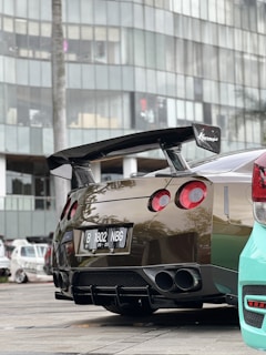 A sports car with a modern design is parked on a paved area near a large building with reflective glass windows. The car features a prominent rear spoiler, dual exhausts, and a glossy finish. Nearby, part of a turquoise vehicle is visible, and other cars are parked in the background.