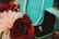 A Tiffany & Co. turquoise gift bag is prominently displayed alongside matching turquoise ring box. The box is open to reveal a sparkling diamond ring with a luxurious setting. Surrounding these items are elegant, red and white artificial flowers, enhancing the luxurious and romantic theme.