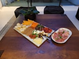 A wooden table displays a charcuterie board with an assortment of cheeses, dried fruits, and meats, along with crackers and fresh herbs. Adjacent to the board, a plate holds thinly sliced cured meats garnished with herbs. A small black pot with a green plant is in the background.