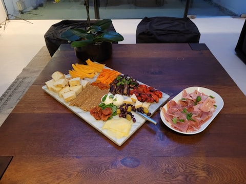 A wooden table displays a charcuterie board with an assortment of cheeses, dried fruits, and meats, along with crackers and fresh herbs. Adjacent to the board, a plate holds thinly sliced cured meats garnished with herbs. A small black pot with a green plant is in the background.