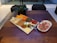 A wooden table displays a charcuterie board with an assortment of cheeses, dried fruits, and meats, along with crackers and fresh herbs. Adjacent to the board, a plate holds thinly sliced cured meats garnished with herbs. A small black pot with a green plant is in the background.