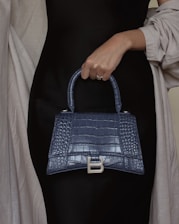 A hand holding a stylish, textured leather handbag with a metallic 'B' clasp. The handbag has a dark blue, crocodile-like pattern and a structured shape. The person is wearing a dark outfit with a light shawl draped over the shoulders.