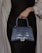 A hand holding a stylish, textured leather handbag with a metallic 'B' clasp. The handbag has a dark blue, crocodile-like pattern and a structured shape. The person is wearing a dark outfit with a light shawl draped over the shoulders.