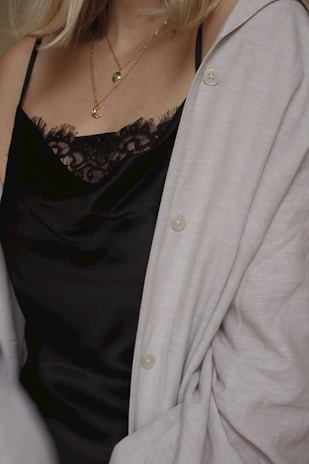 A close-up shot of a person wearing a black lace-trimmed camisole. They have layered gold necklaces and a light-colored cardigan draped over the shoulders.