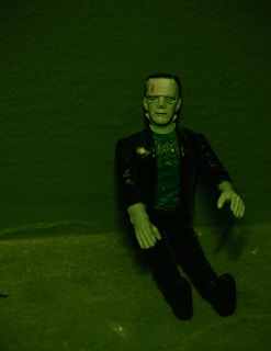 A green-toned image of a Frankenstein's monster figurine sitting against a textured background. The figure has a flat top head with bolts on its neck, detailed facial features, and wears a dark jacket over a textured green top. Its arms are posed slightly bent at the elbows.