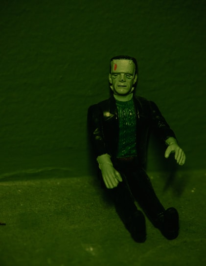 A green-toned image of a Frankenstein's monster figurine sitting against a textured background. The figure has a flat top head with bolts on its neck, detailed facial features, and wears a dark jacket over a textured green top. Its arms are posed slightly bent at the elbows.